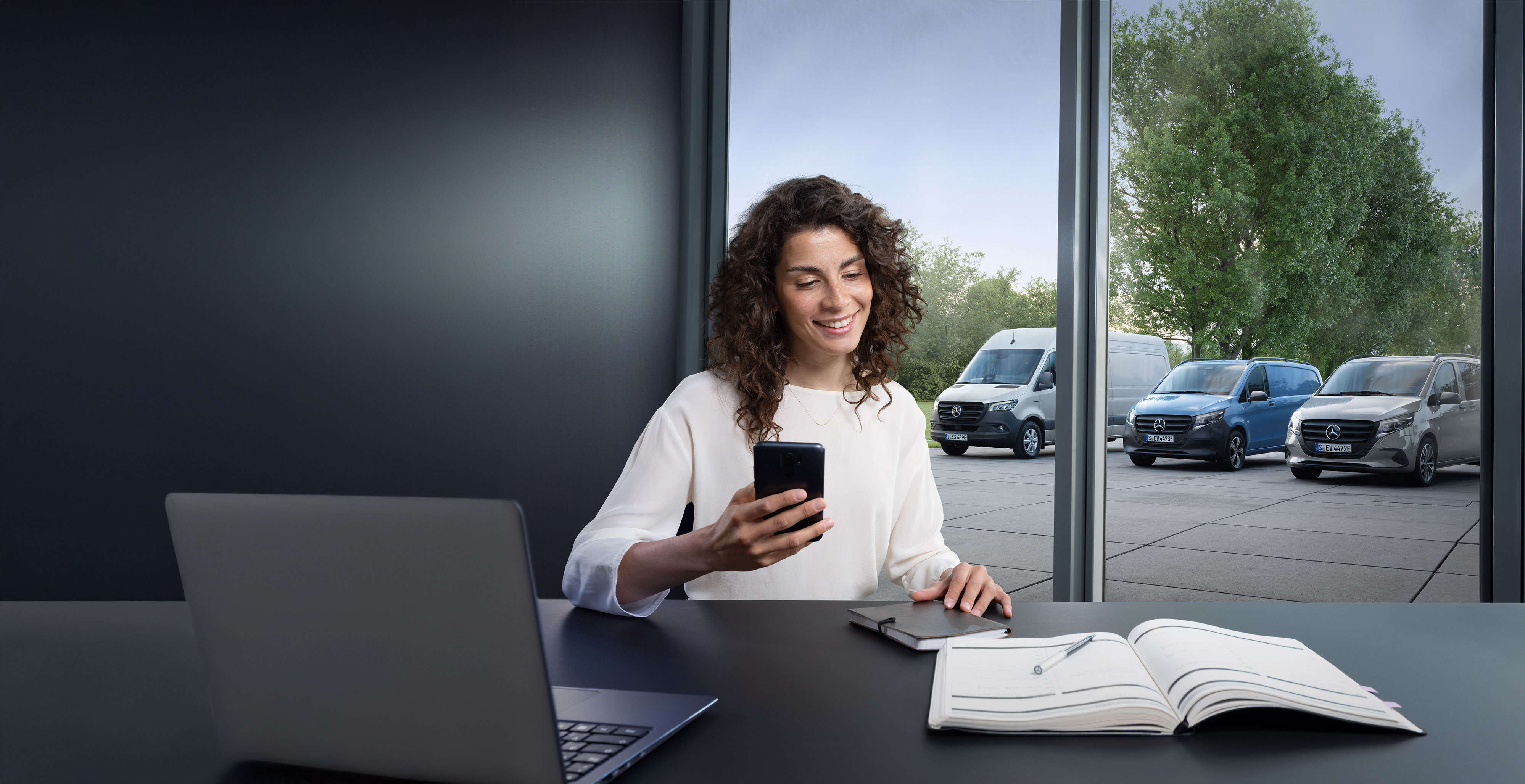 Mercedes-Benz | Transporterbetreuung Frau mit Smartphone an einem Schreibtisch, im Hintergrund mehrere Mercedes-Benz Transporter – fachkundige Unterstützung im Bereich Business Care.